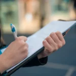 Person holding a pen and writing on a clipboard with papers, wearing a blue garment and dark jacket, with a blurred background