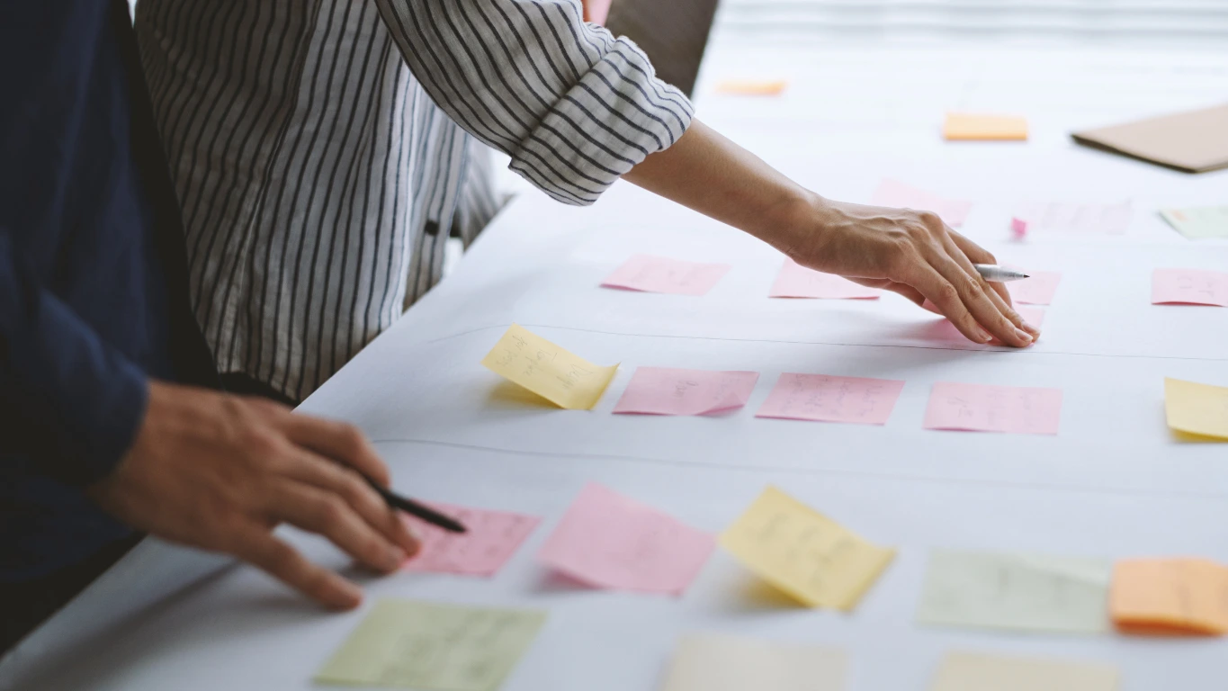 Close-up of two people arranging colorful sticky notes on a large white table, with one person wearing a striped shirt and another in a dark long-sleeve shirt.